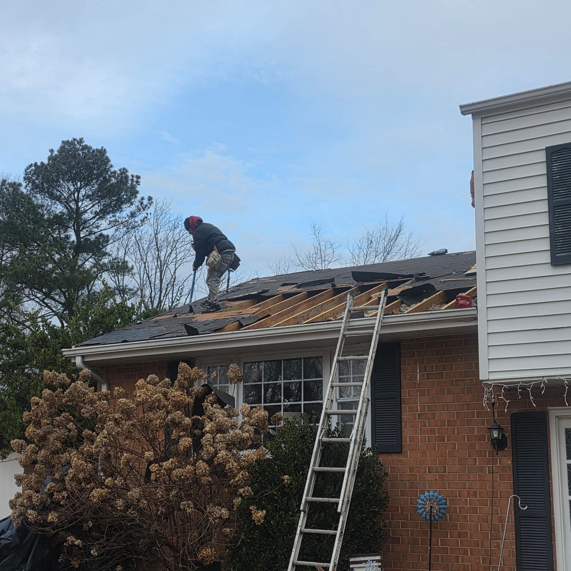 Roofer on a partially torn-off roof. Ladder leans against the house, cloudy sky in the background.