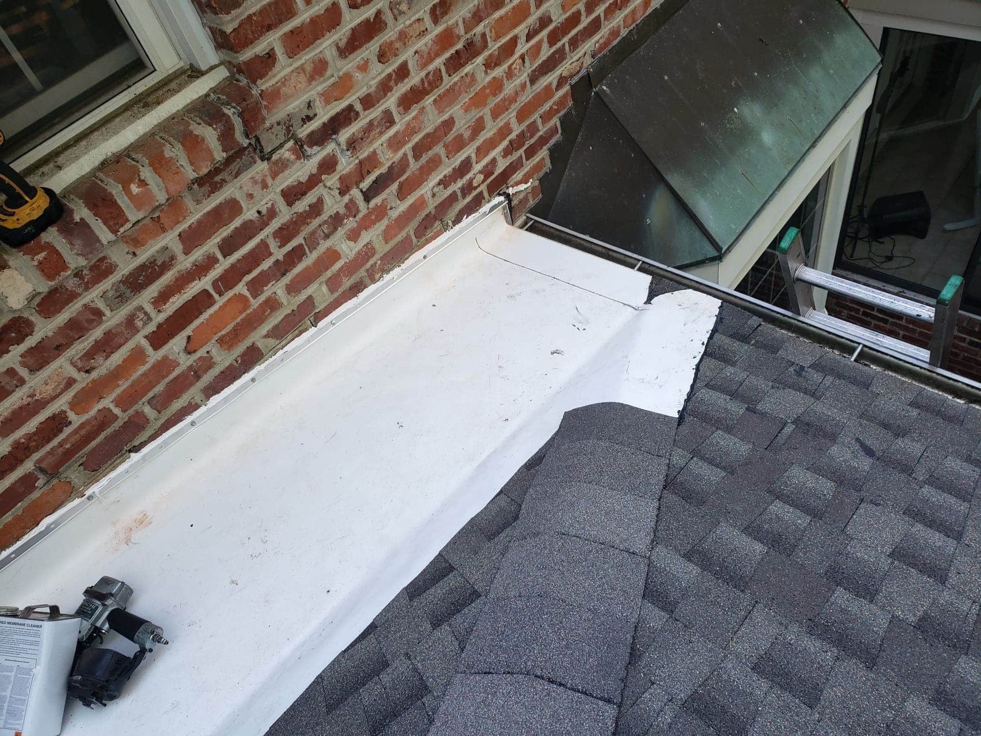 White flashing installed on a roof next to a brick wall. A nail gun is visible.