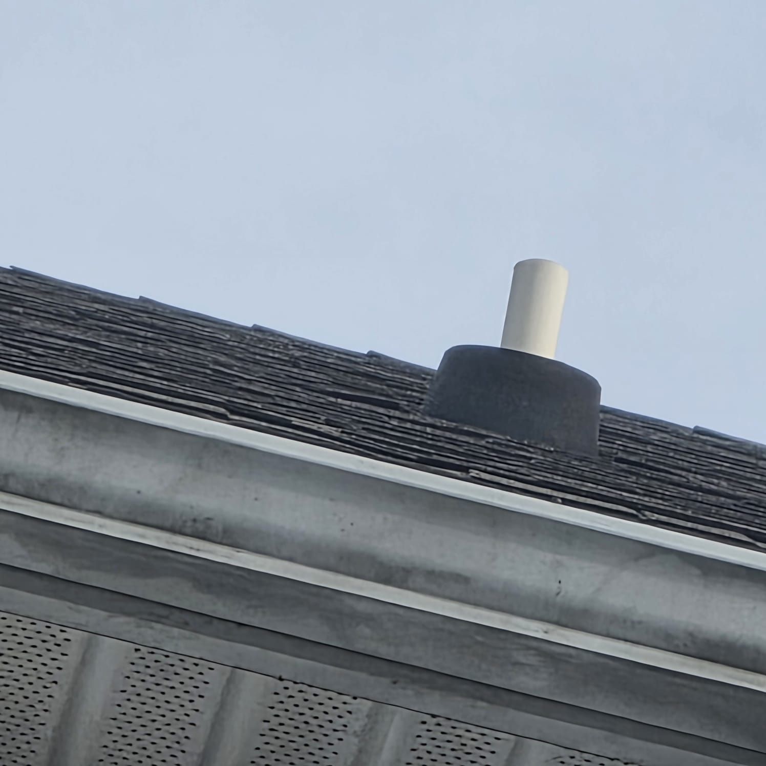 Roof with gutter and vent pipe. Black shingles, gray gutter, white vent, and a blue sky.