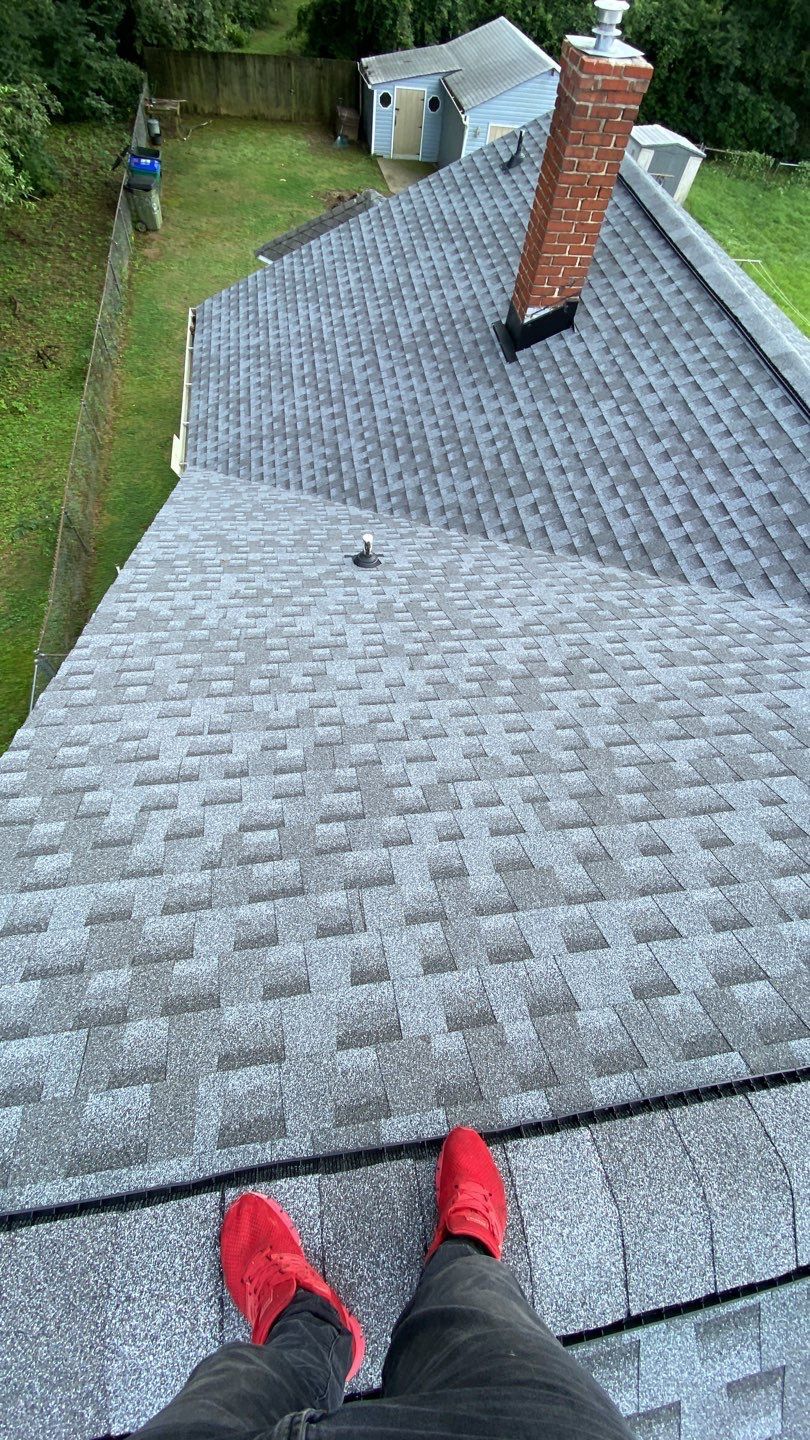 Person in red shoes standing on a gray shingle roof. Brick chimney visible; background includes grass and a small building.