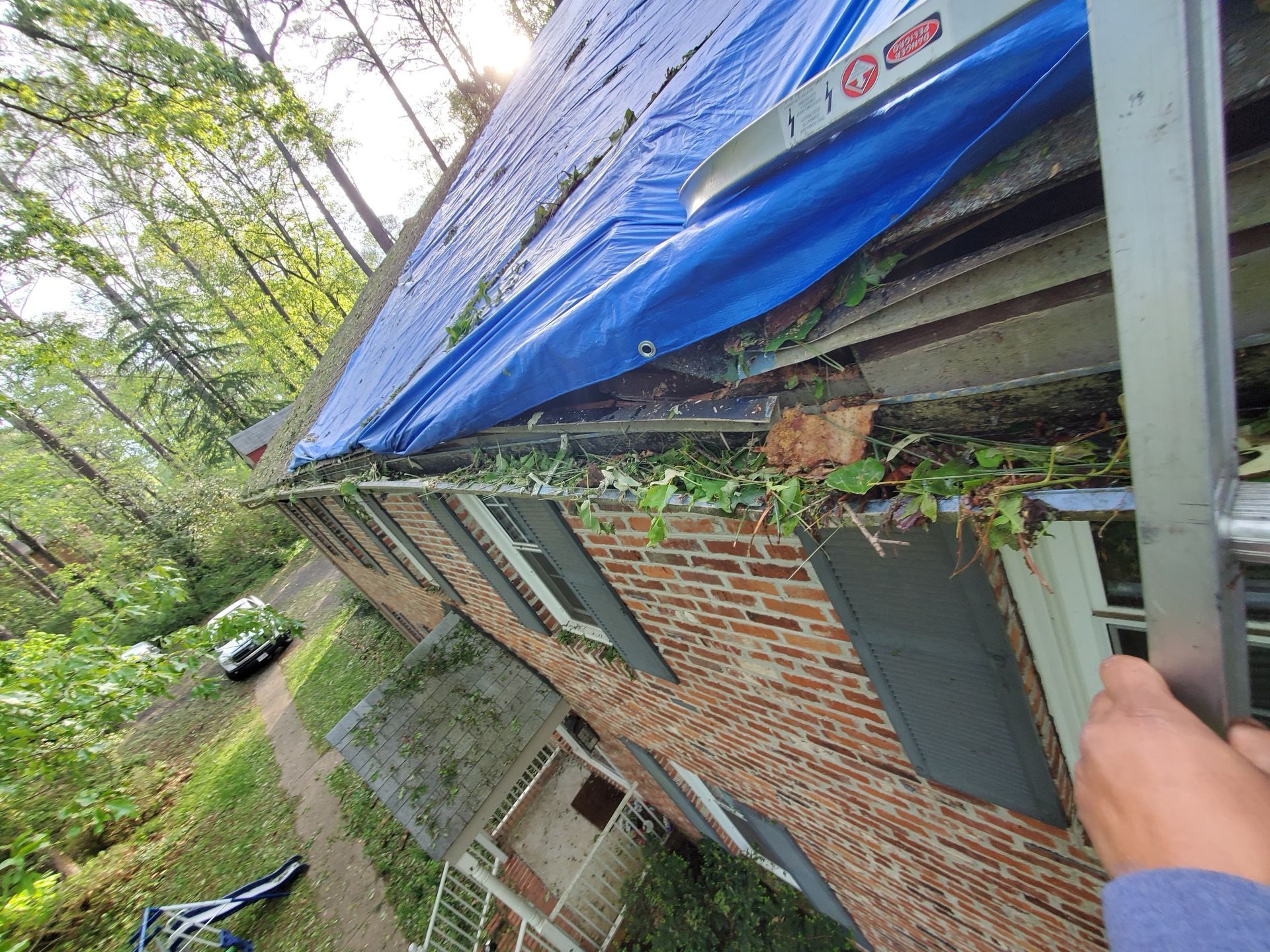 House with blue tarp on roof, ladder, and debris-filled gutters.