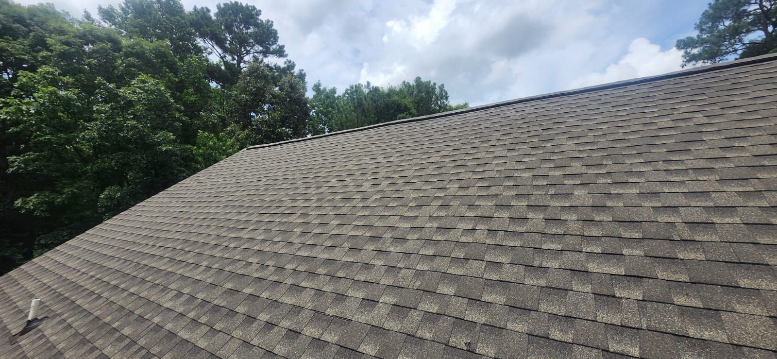 Overhead view of a weathered asphalt shingle roof, with trees and a cloudy sky in the background.