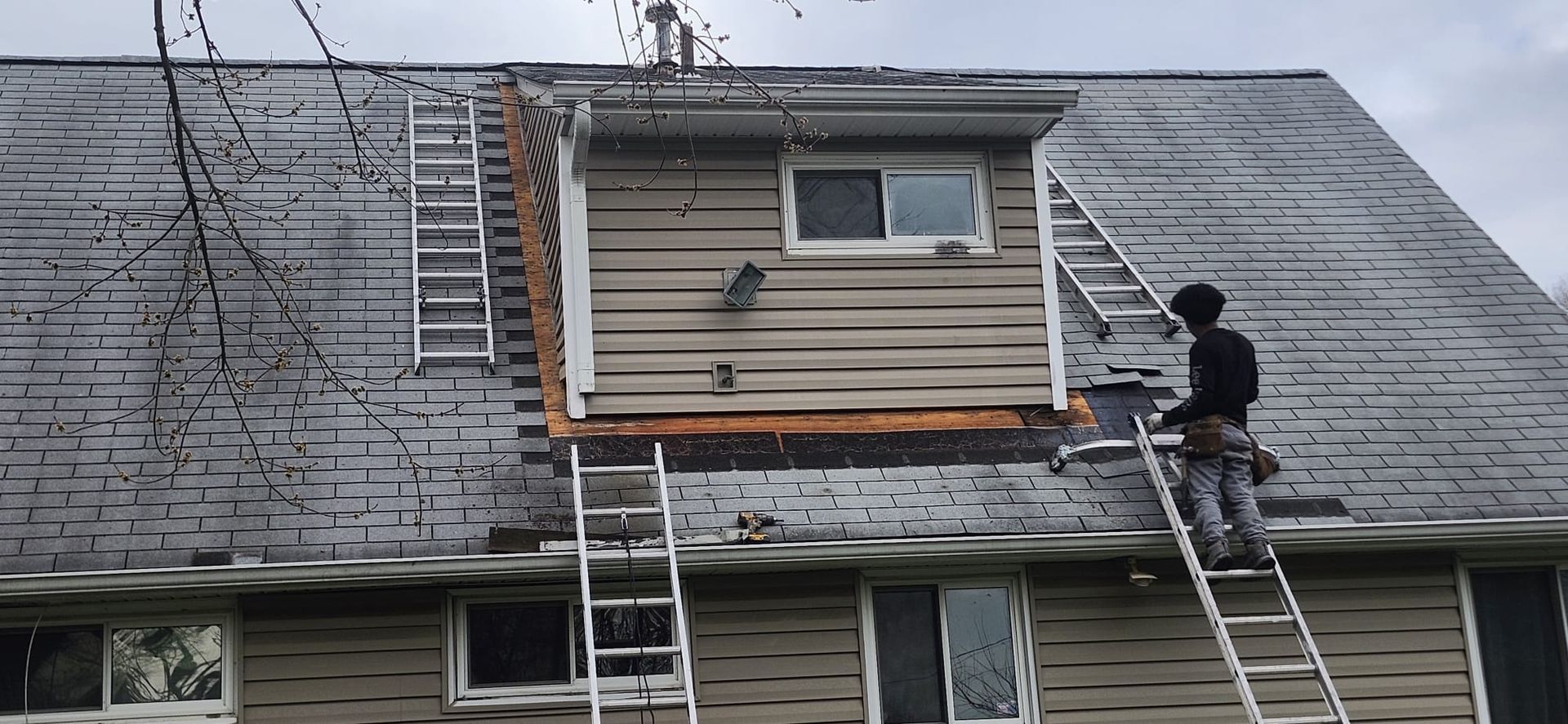 Person on a ladder repairing a roof on a building. There is another ladder and a dormer. Cloudy sky.
