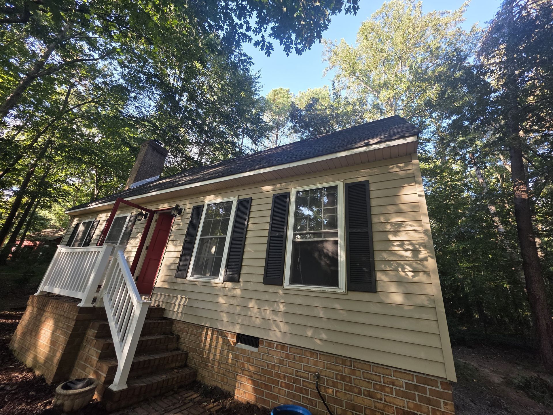 Tan house with black shutters, red door, and brick foundation, surrounded by trees.