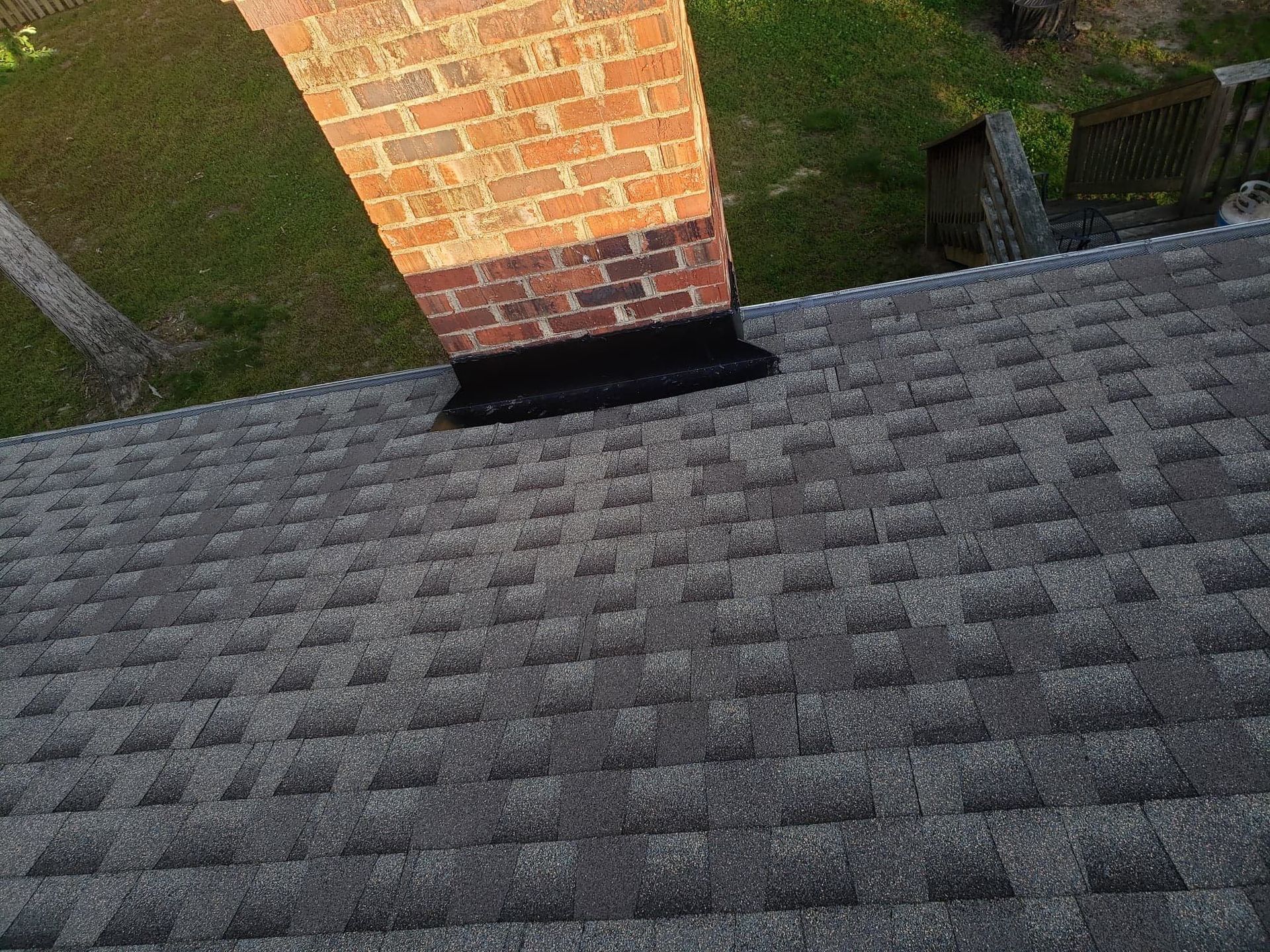 Brick chimney on a gray shingle roof, surrounded by green grass and a tree.