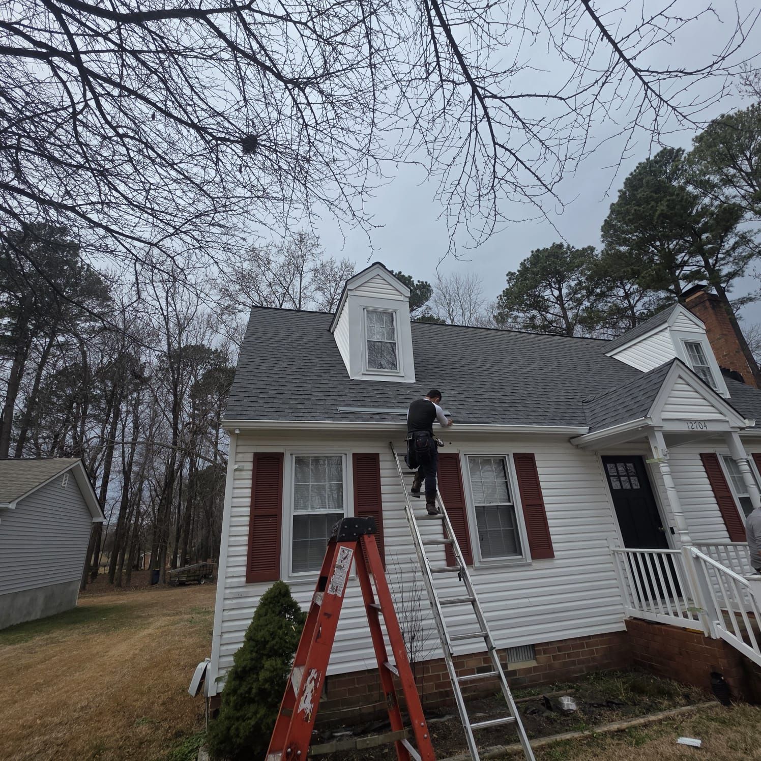 Person on a ladder cleaning gutters of a white house with red shutters on a cloudy day.