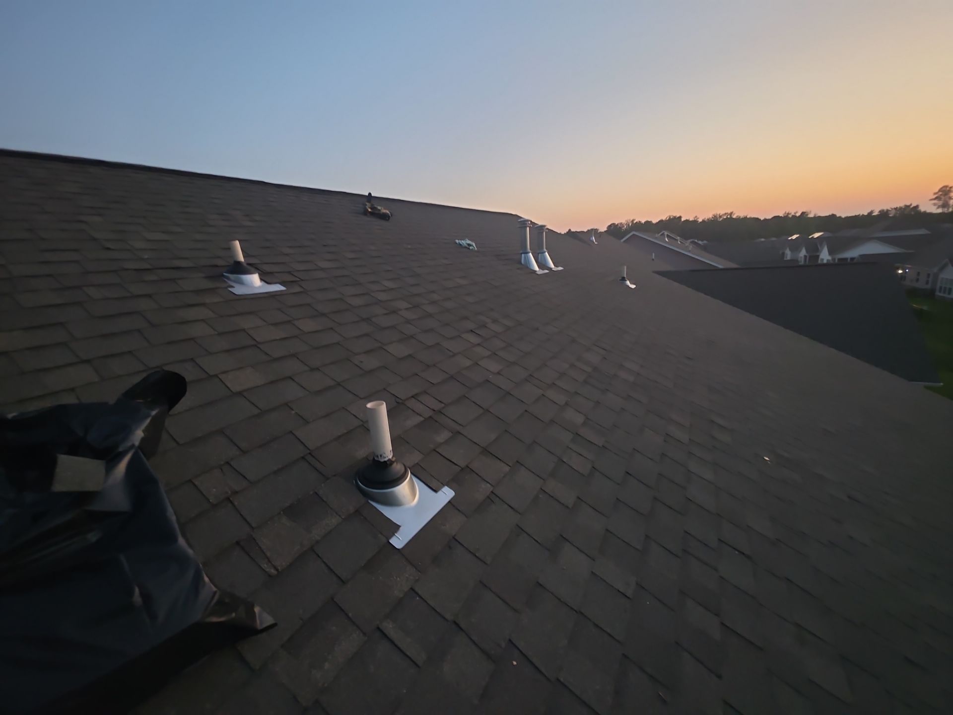 Shingled rooftop at sunset with several vent pipes and a dark tarp in the foreground.