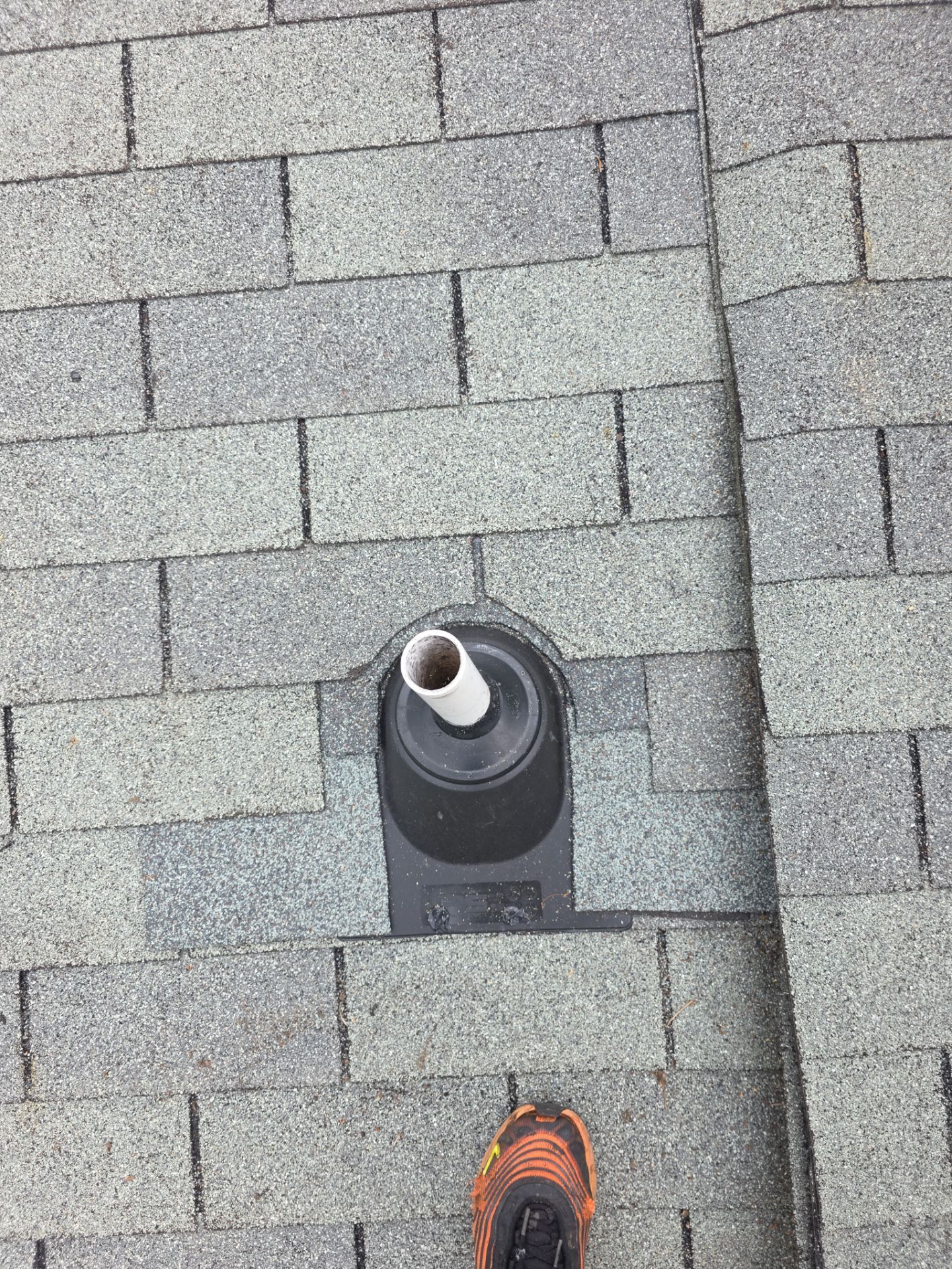 View from a roof edge: a vent pipe on gray shingles with an orange shoe at the bottom.