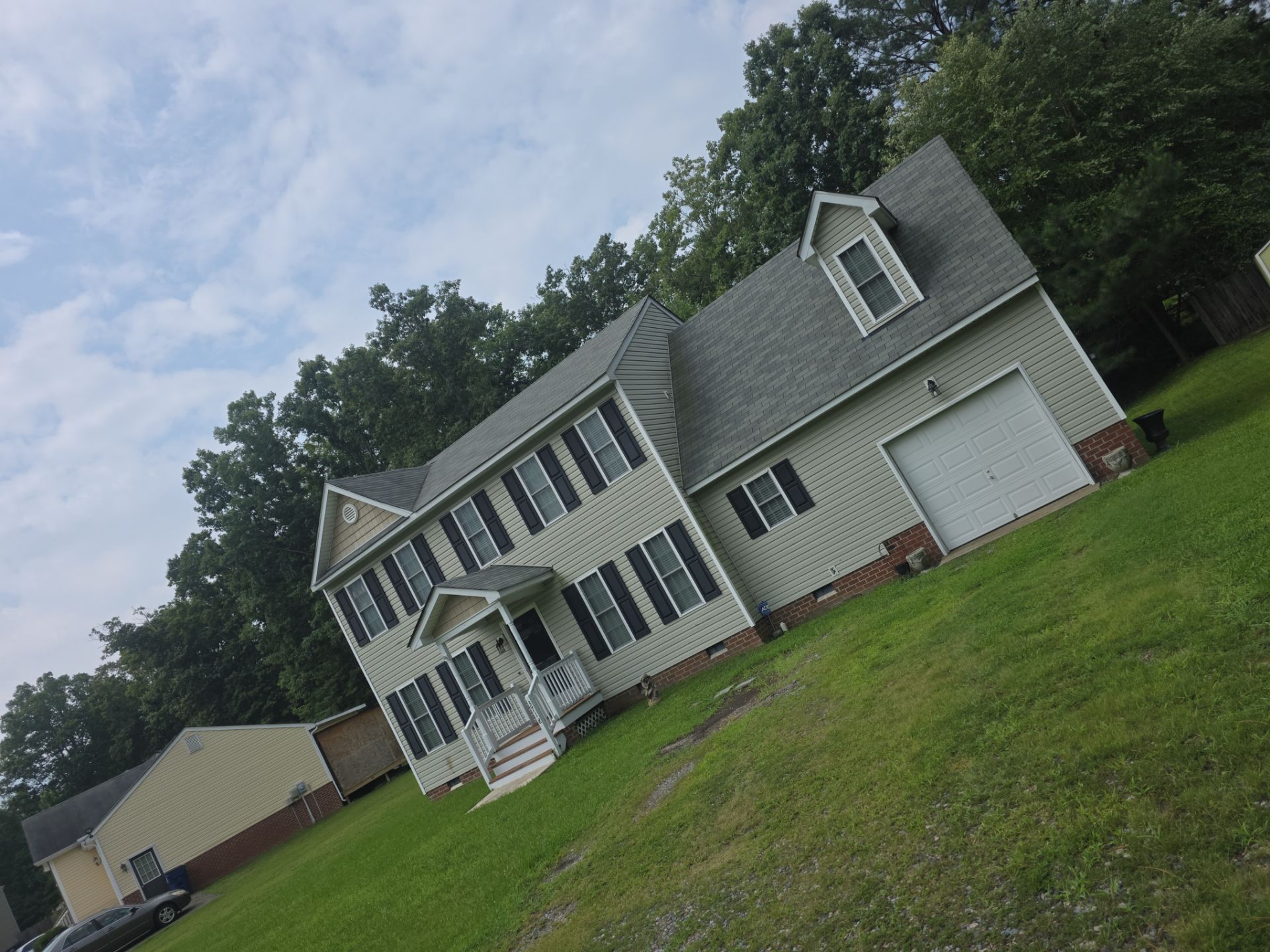 Tilted view of a gray two-story house with white trim, a garage, and a large green lawn under a cloudy sky