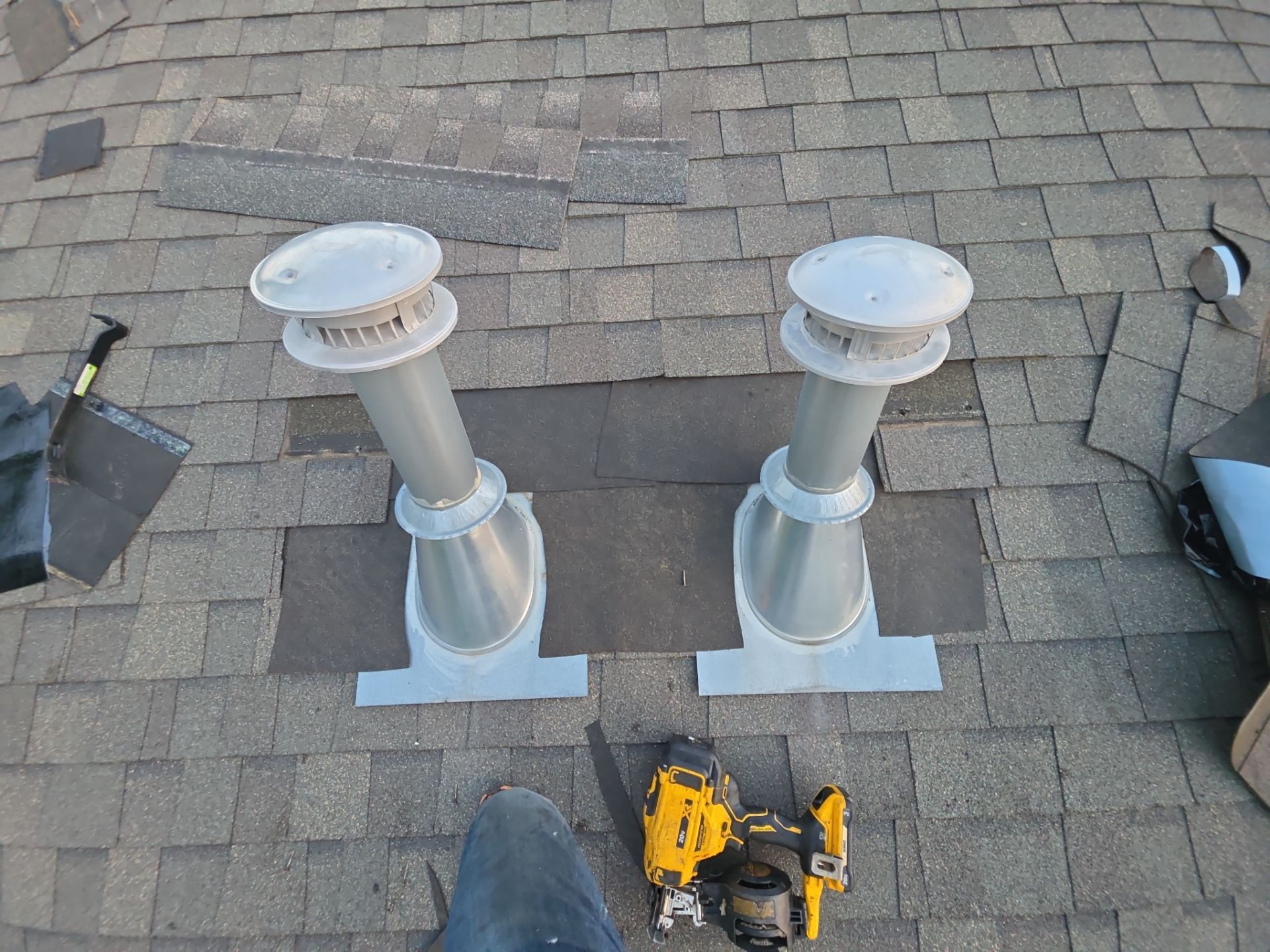 Two white roof vents on shingles, with a yellow tool sitting below them.