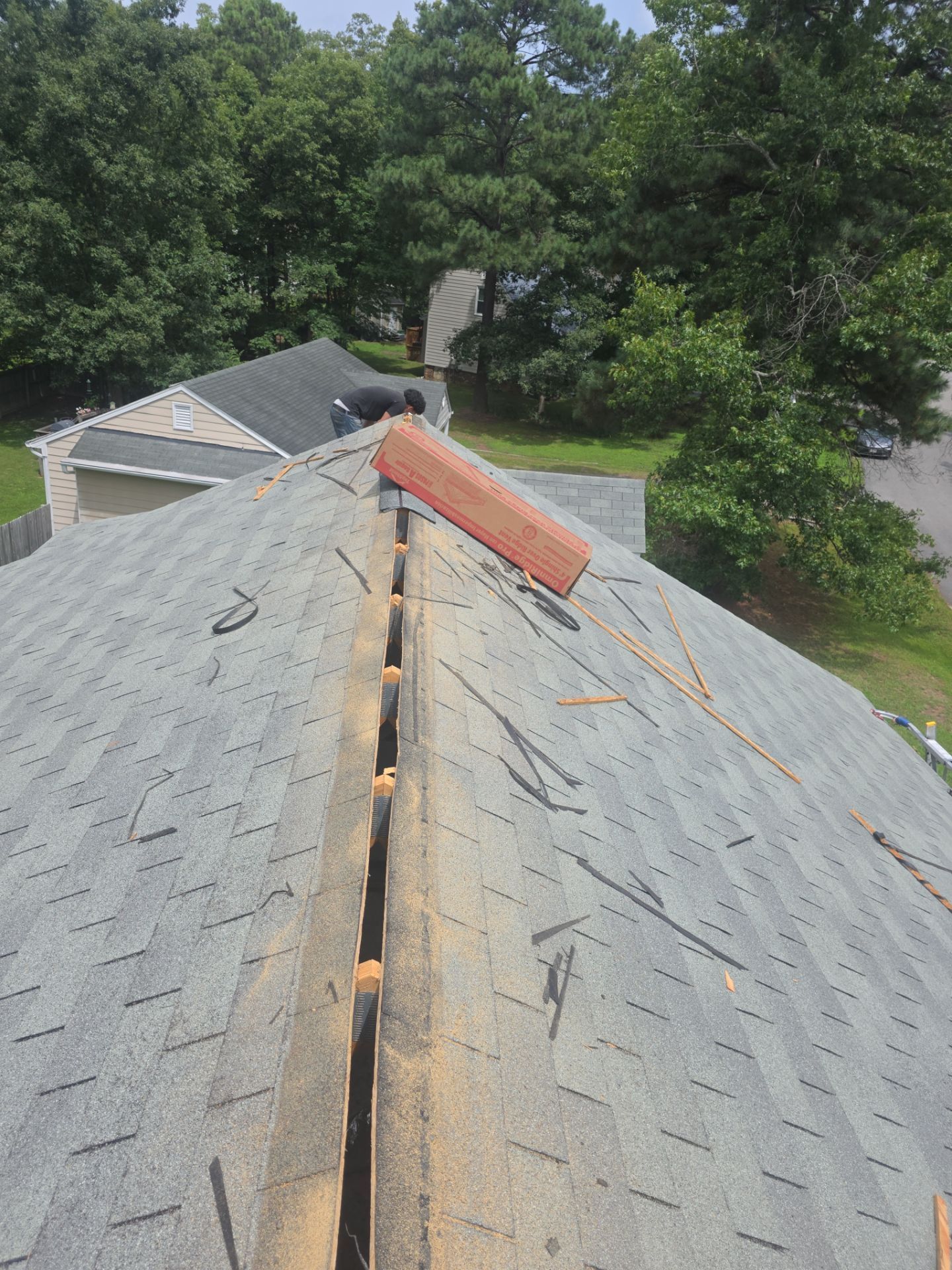 Roof with damaged shingles and exposed ridge, overlooking trees and a neighboring house.