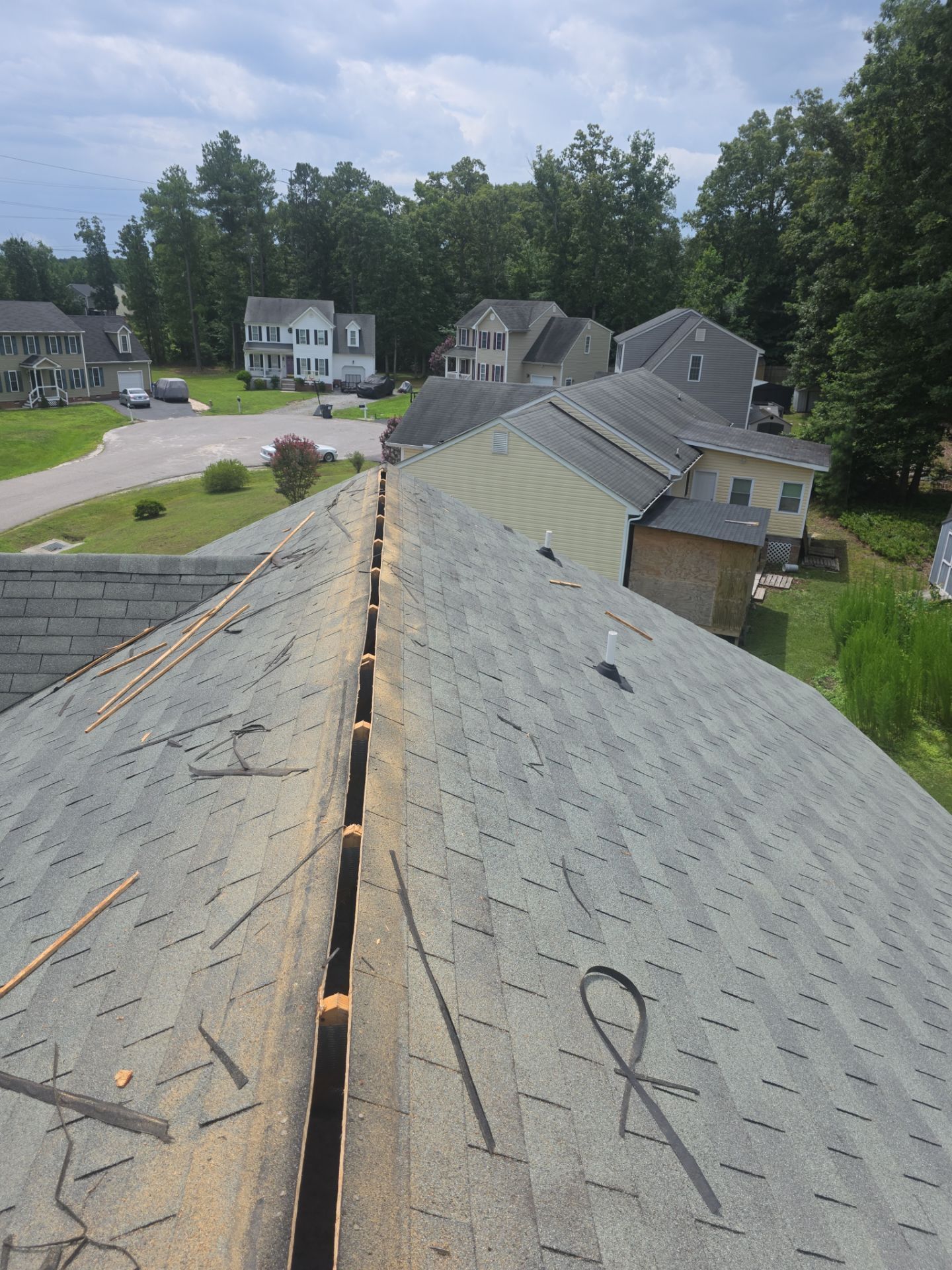 Roof ridge with damaged shingles and gaps, viewed from above in a suburban neighborhood.