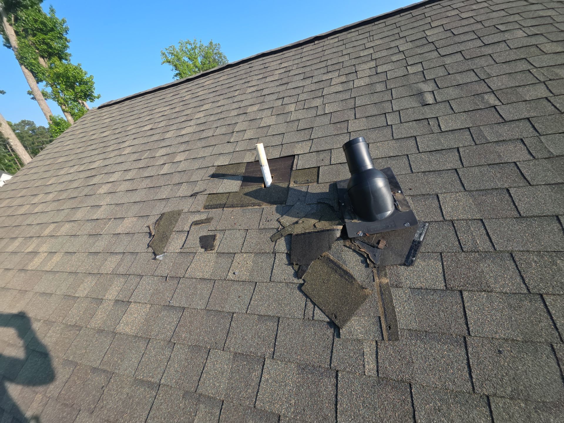 Damaged shingle roof with a black vent pipe and exposed underlayment near a roof repair area