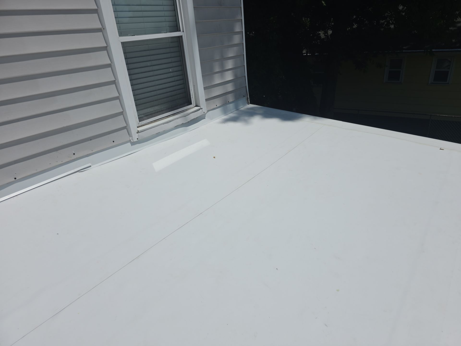 White rooftop or deck beside gray house siding and a window, with a dark building in the background