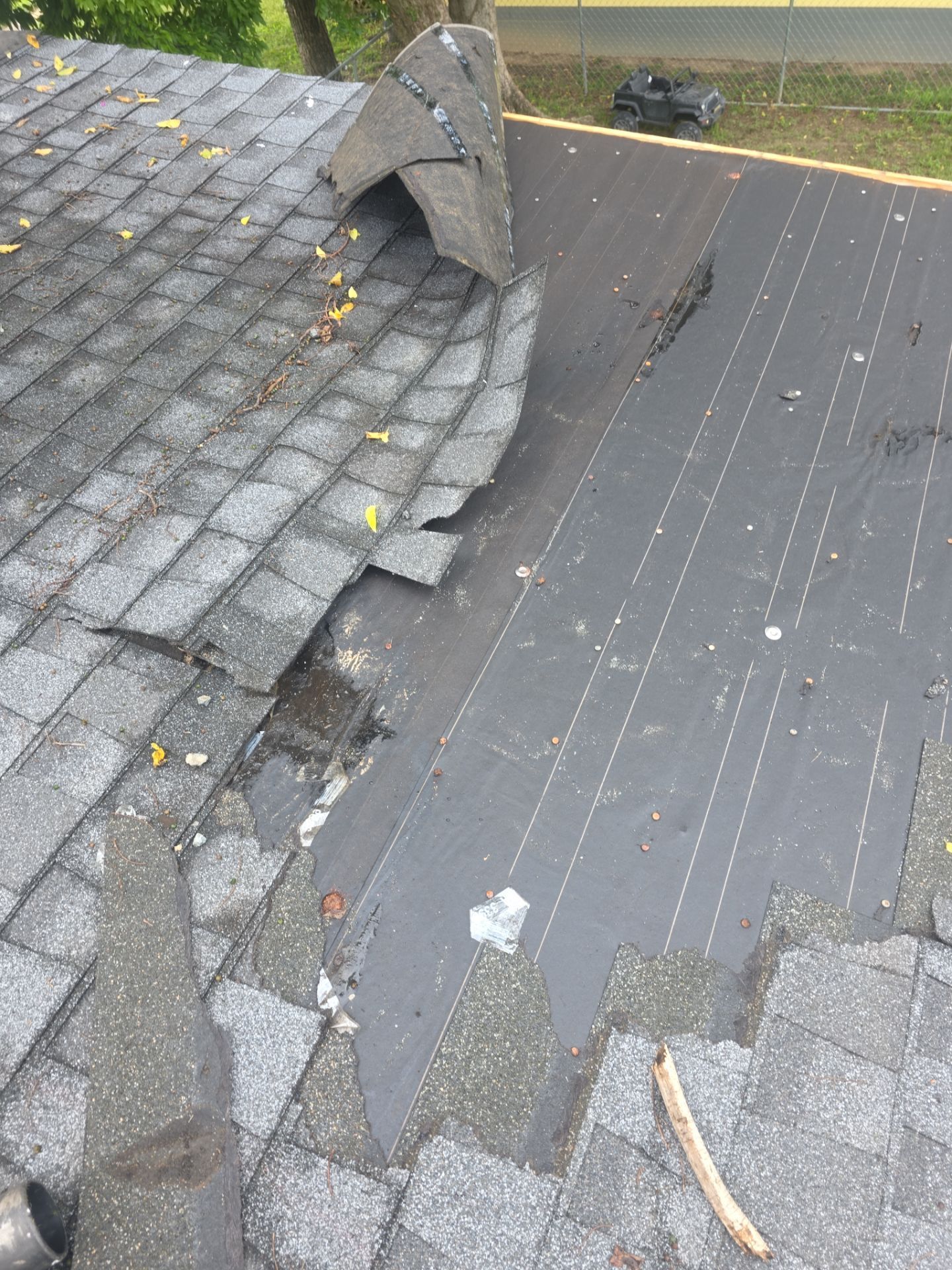 Damaged roof with missing shingles and exposed underlayment beside intact gray shingles