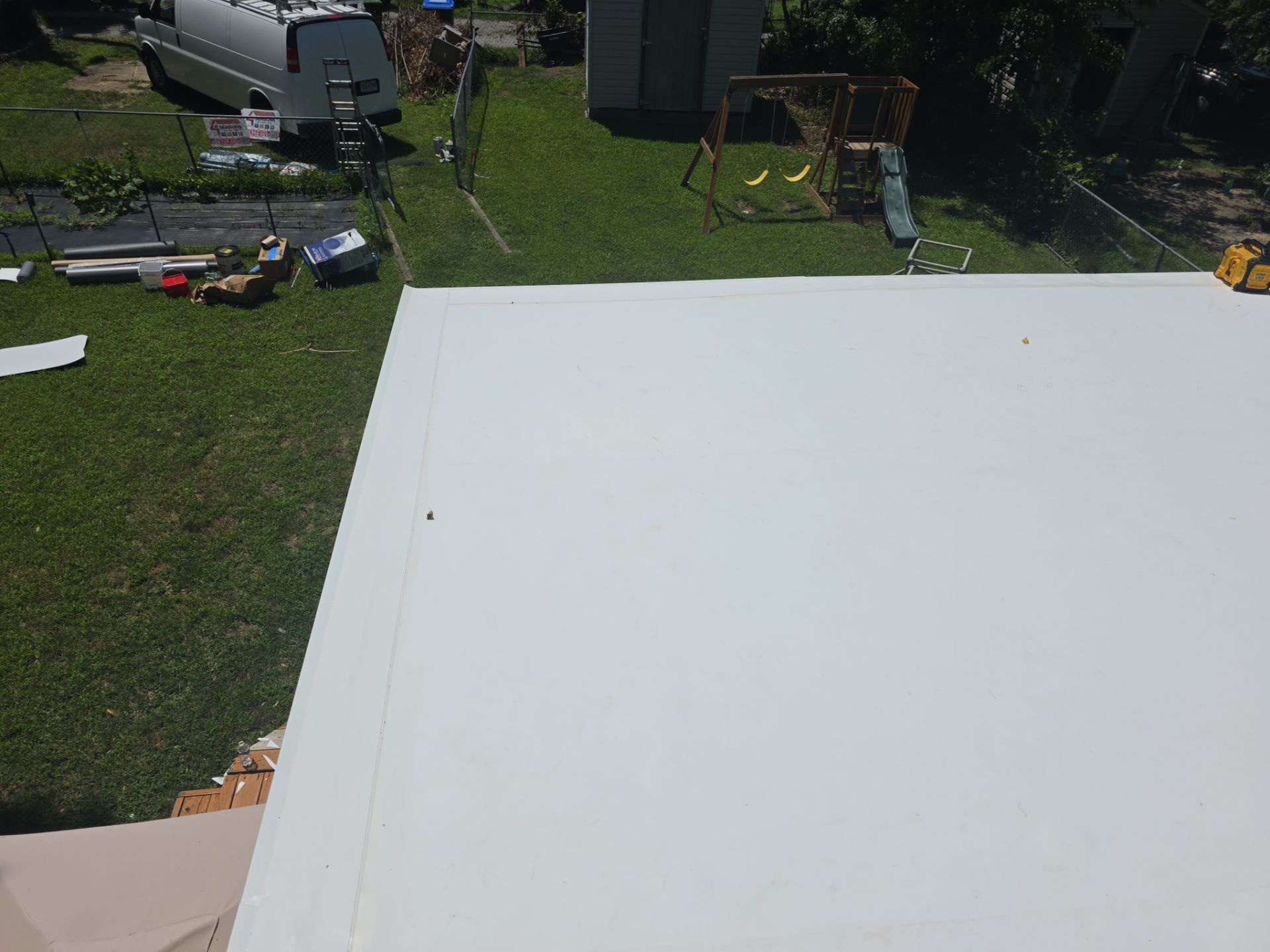 White rooftop or tarp in foreground with a grassy yard, trees, and scattered outdoor equipment in the background