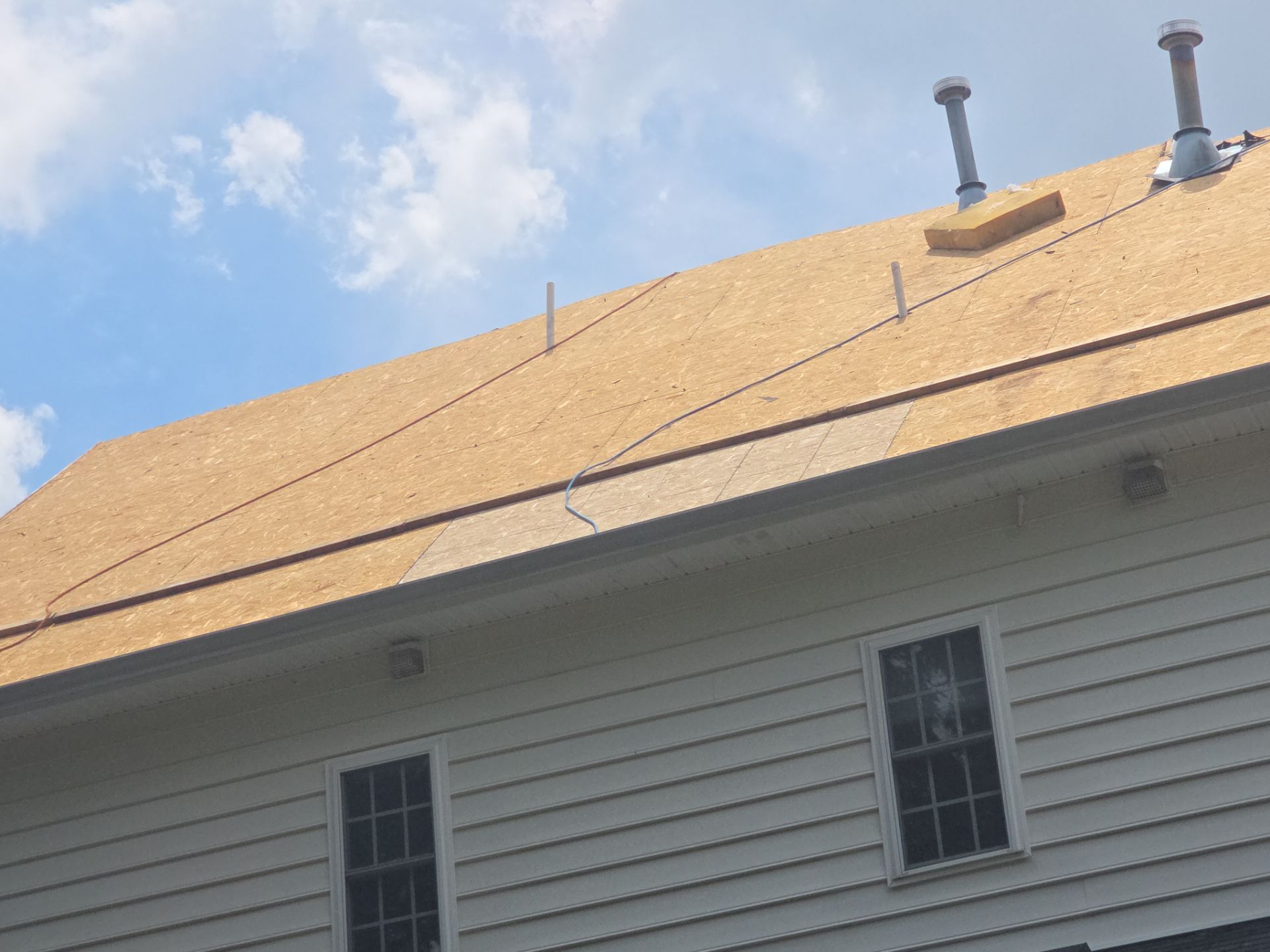 House roof under construction with plywood sheathing, vents, and white siding against a blue sky