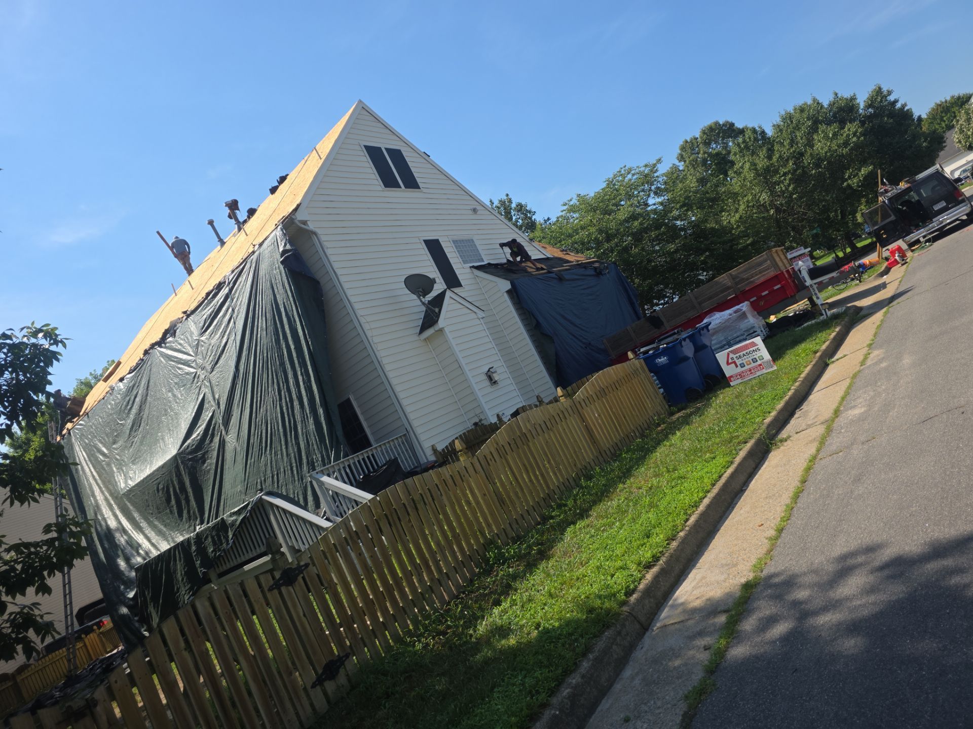 Damaged house with tarp-covered roof beside a fenced sidewalk on a sunny street