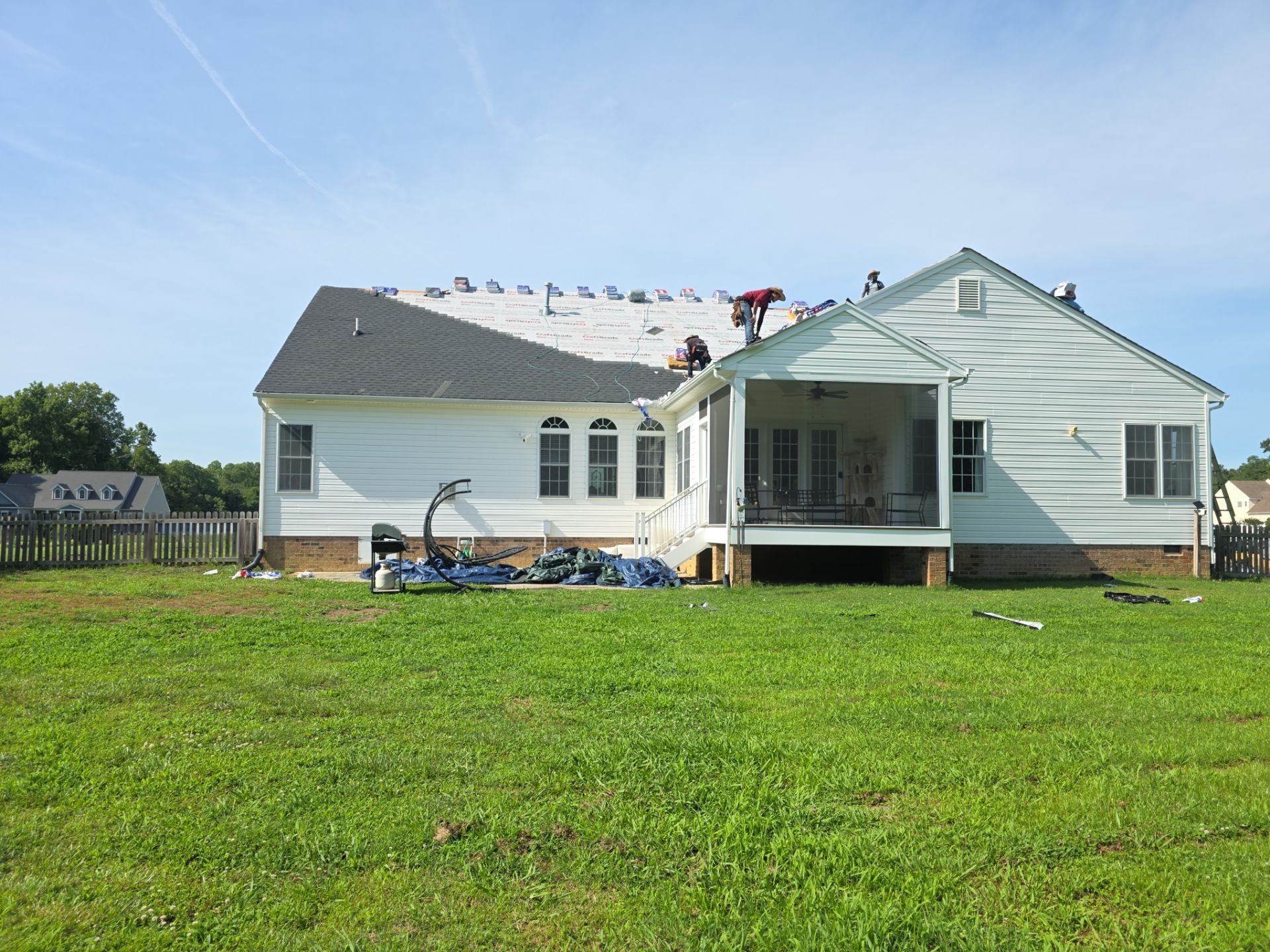 White house with a covered porch on a grassy lawn, roof damaged and cluttered with debris under a blue sky