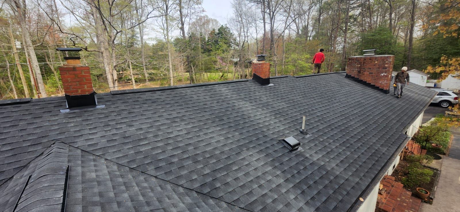 Black shingled roof with brick chimneys and a worker inspecting it near a wooded yard