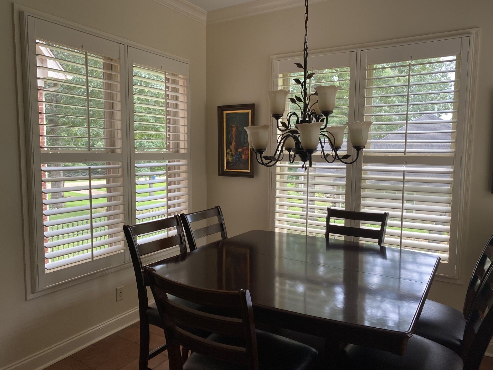A dining room with a dark wood table, four chairs, and a chandelier. Two windows with white shutters let in natural light.