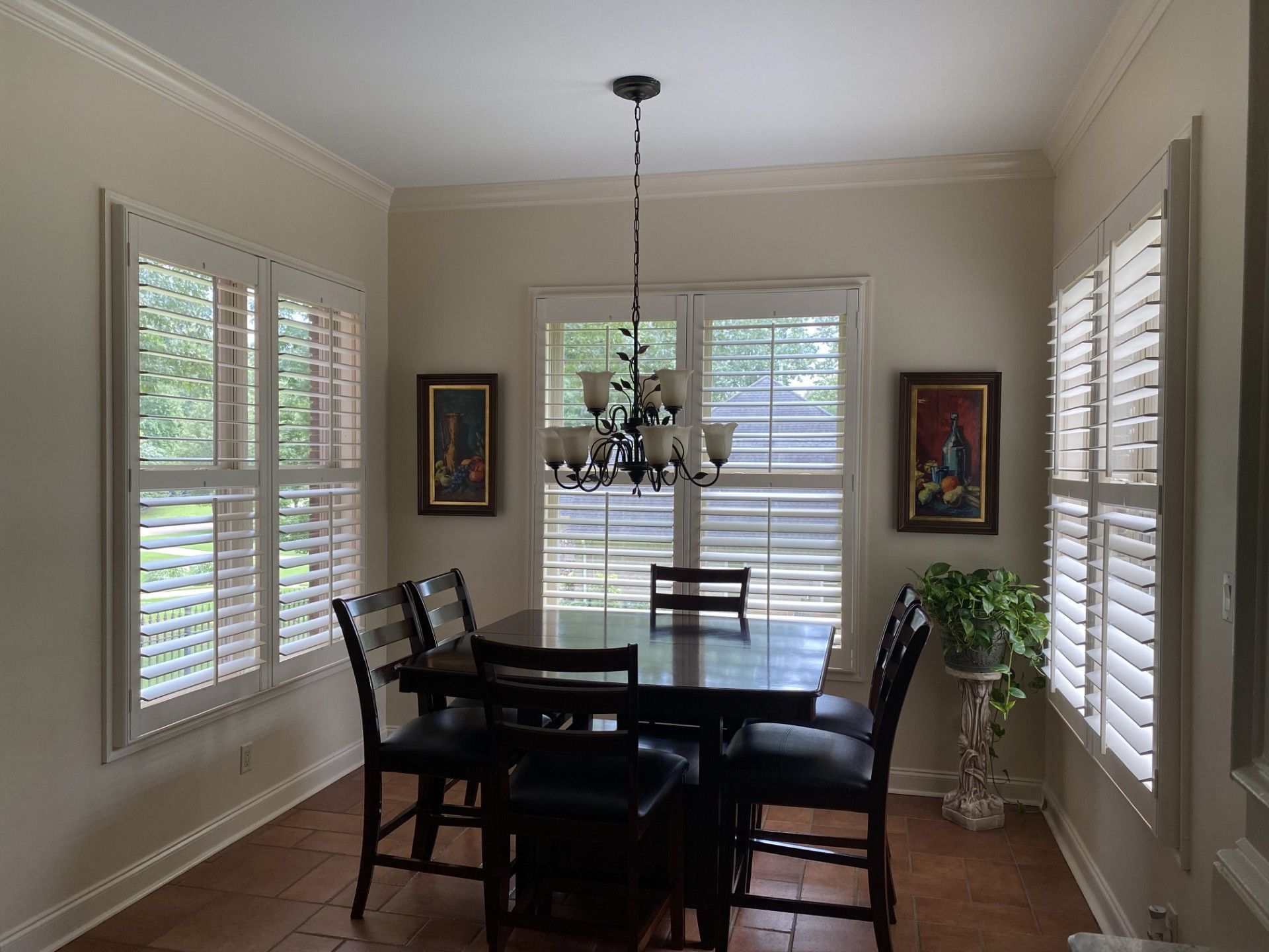 Dining room with a dark wood table and chairs, under a chandelier. Windows with white shutters flank the table, with two paintings on the walls.