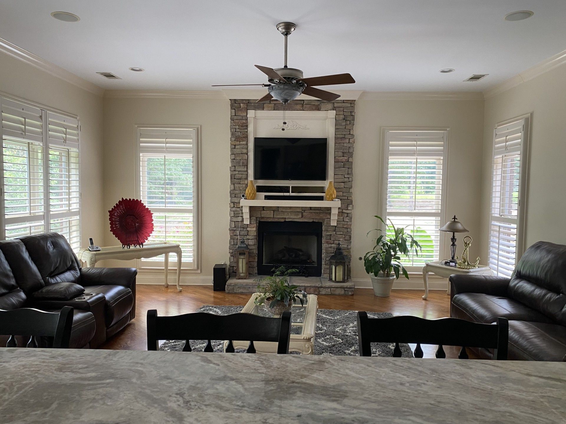 A living room with a stone fireplace, two black leather couches, and large windows with blinds.