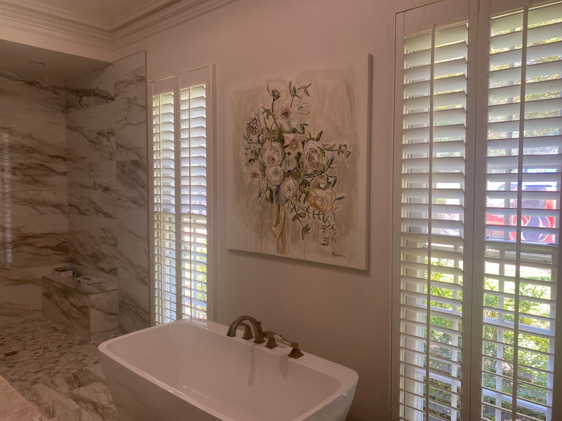 A modern bathroom with a white freestanding tub, marble shower, floral art, and white shutters on windows.