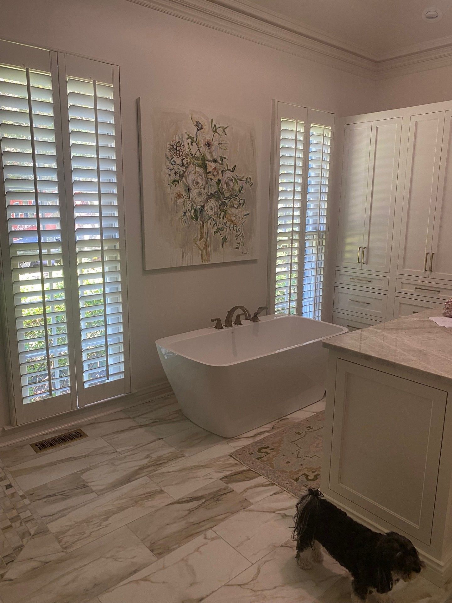 Bathroom with white shutters, freestanding tub, marble tile floor, artwork, and a small dog.