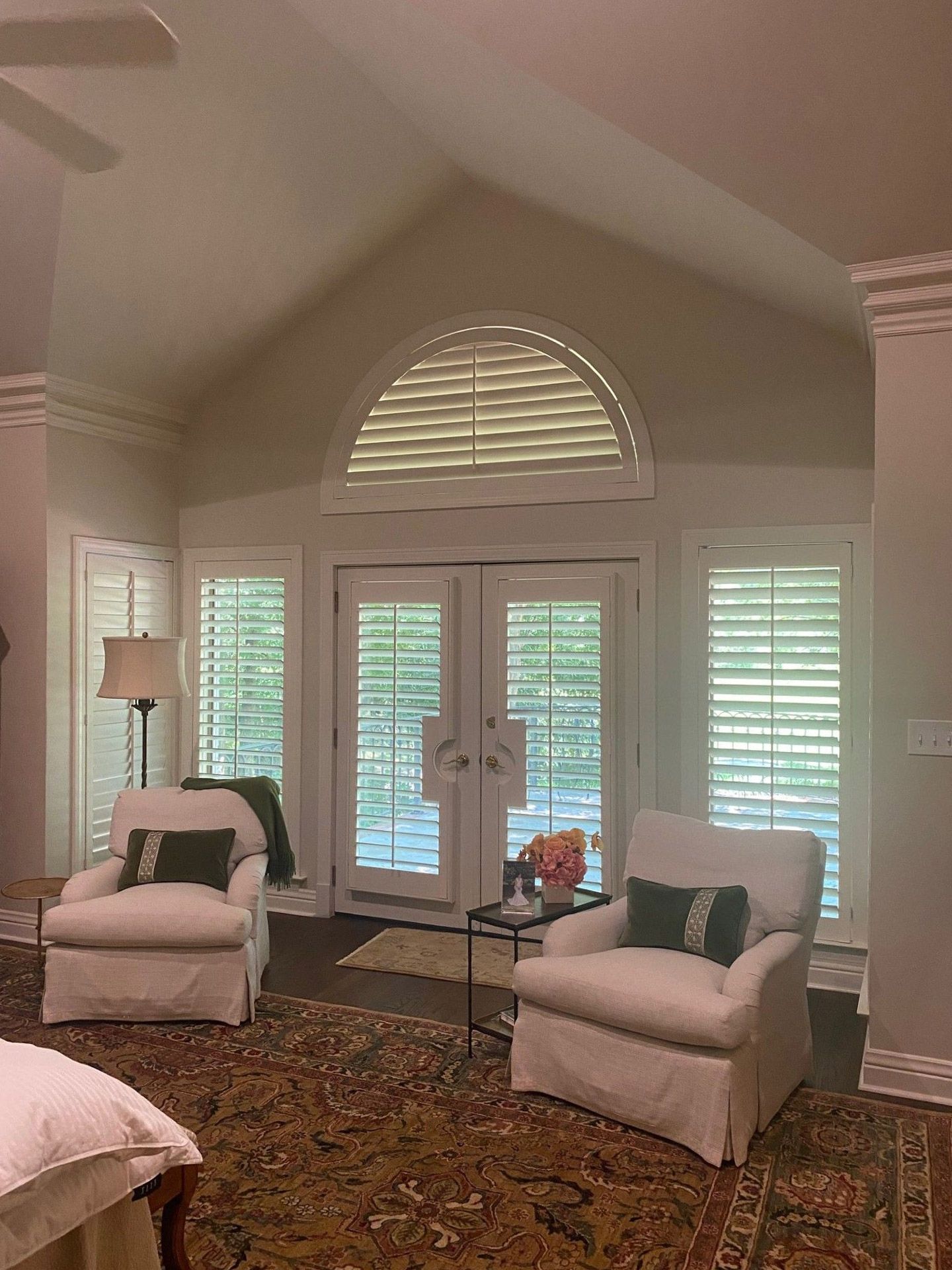 Bedroom with two white armchairs, an area rug, and French doors with shutters. A domed window above the doors also has shutters.
