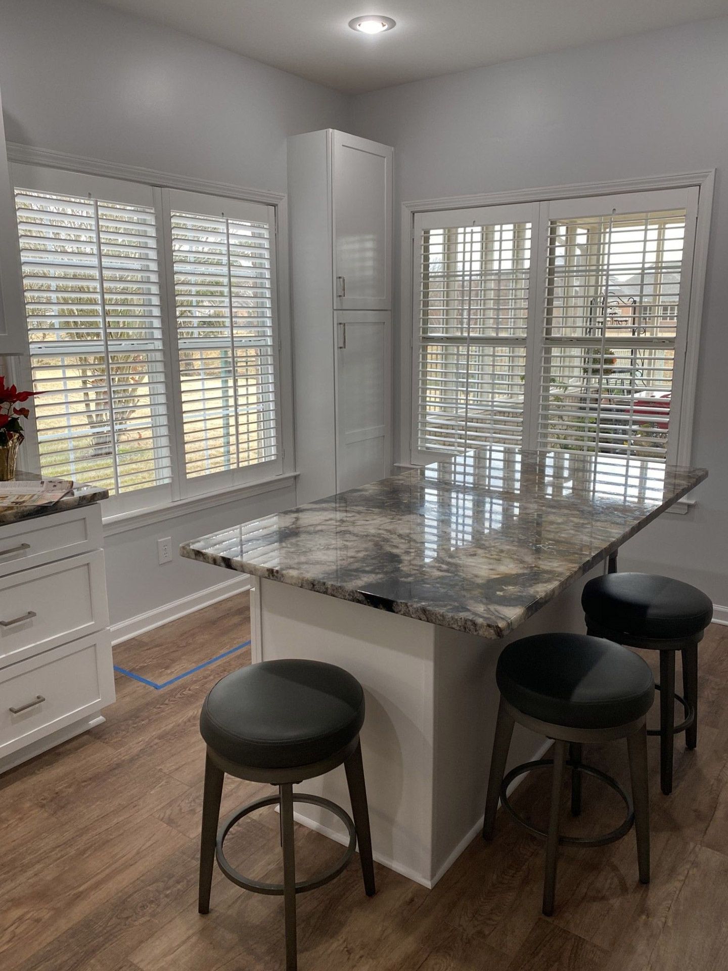 A kitchen with a granite-topped island and bar stools. White cabinets, windows with blinds, and wood-look flooring are also present.