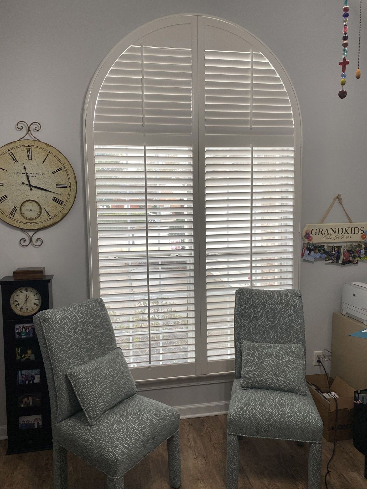 Interior view with arched window, white shutters, two patterned chairs, clock, and decorations.