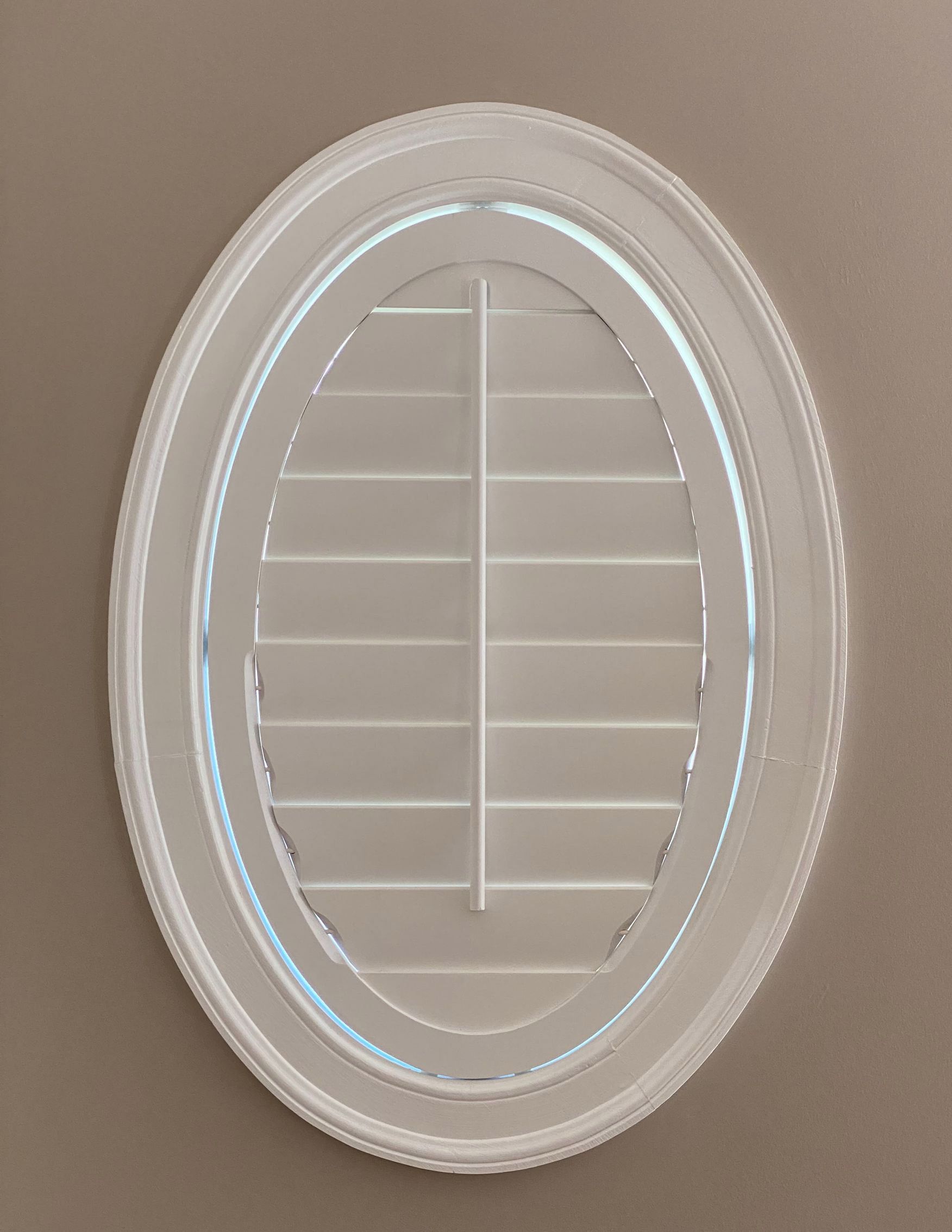 Oval window with white shutters and a white trim against a tan wall.