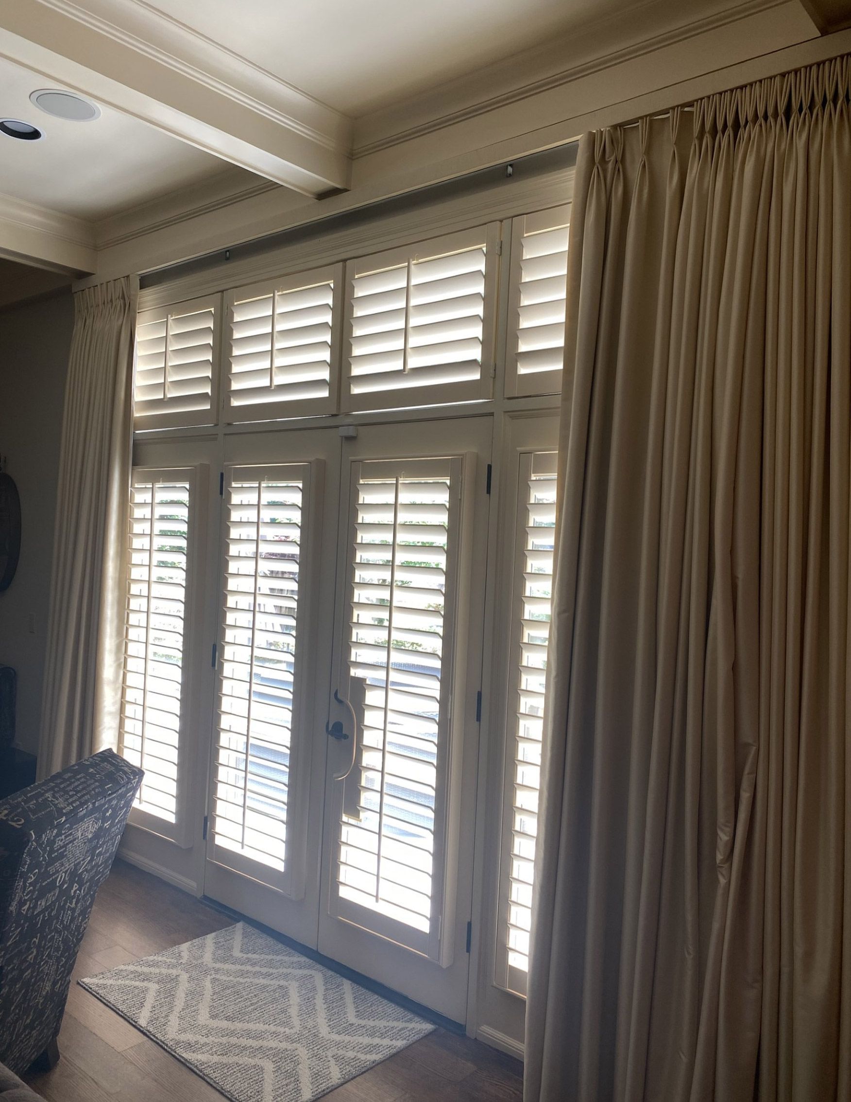 Interior view of French doors with white shutters and beige curtains. A patterned rug is in front of the doors.