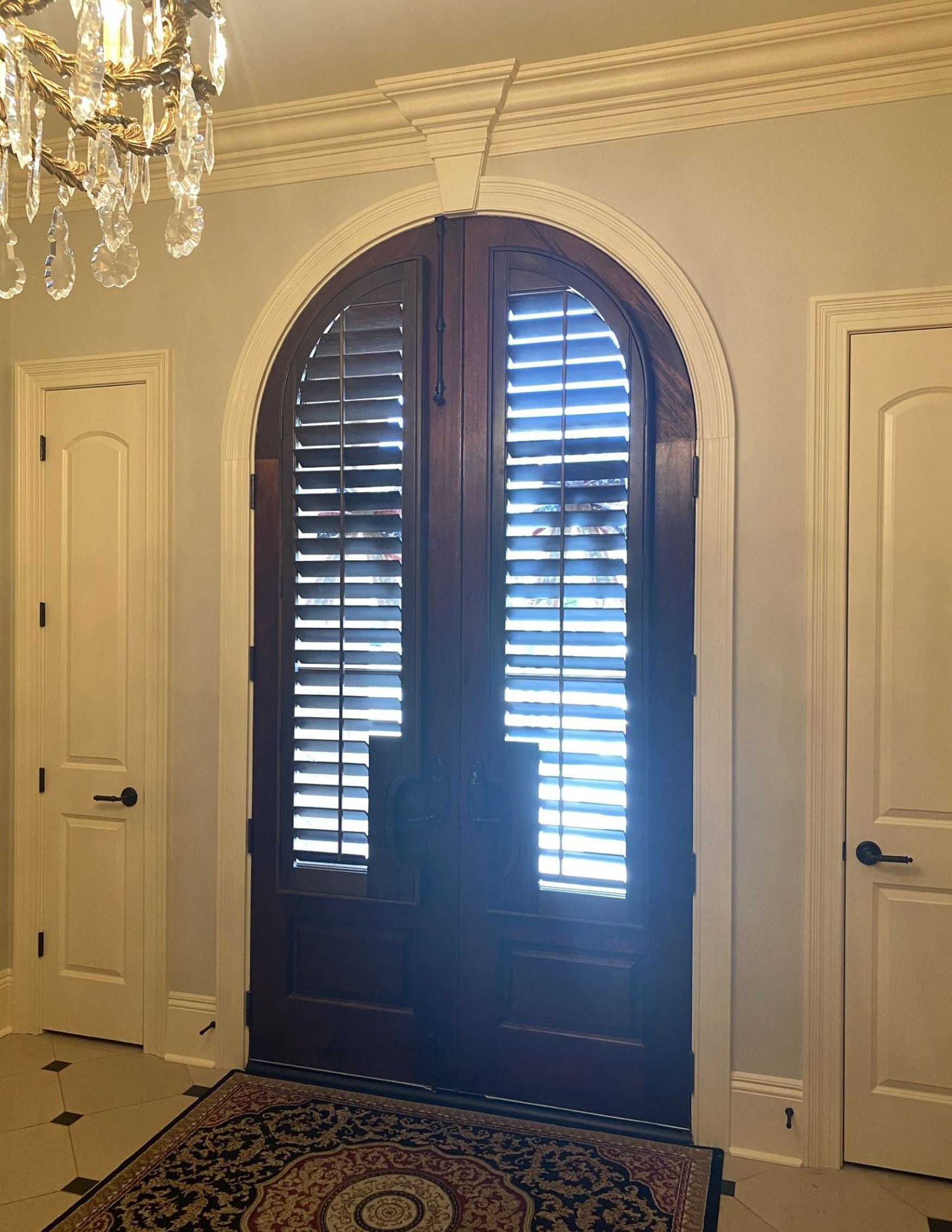 A doorway with dark wood, arched double doors and closed shutters. There are white doors, trim, and a chandelier in the room.