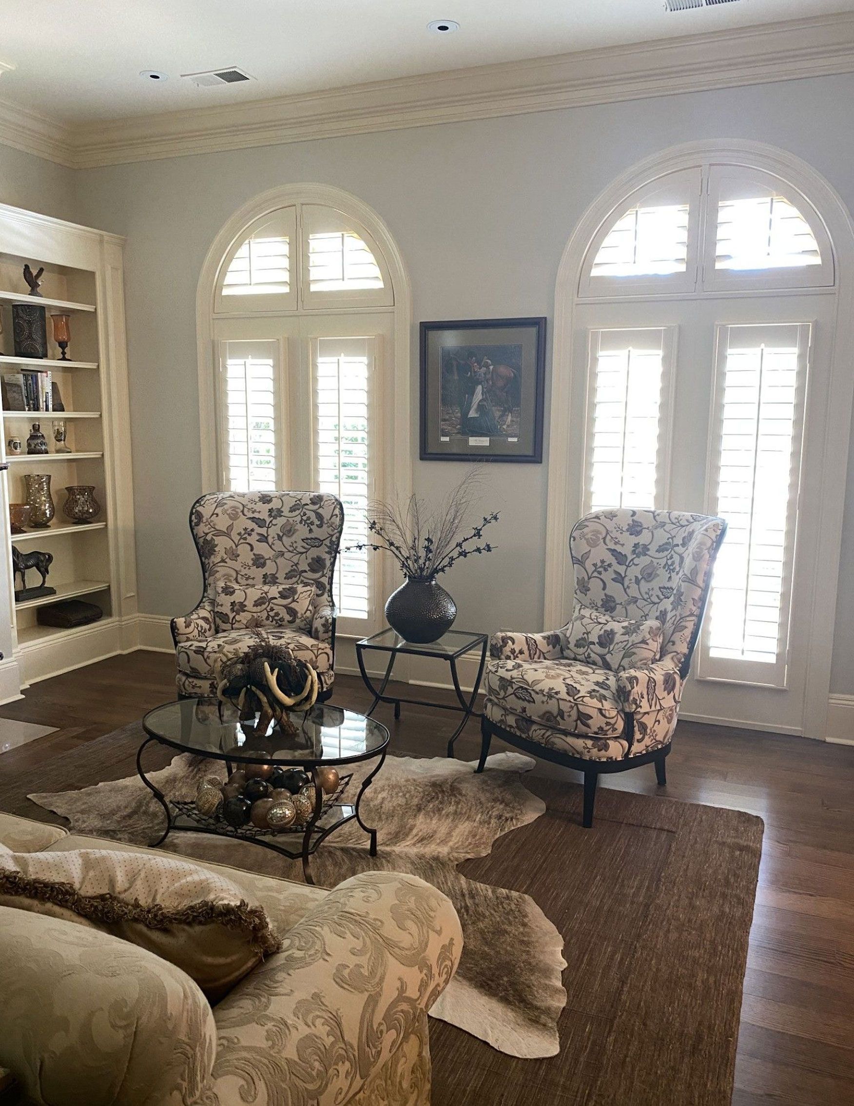 Elegant living room with two floral armchairs, a round glass table, and arched windows with shutters. A built-in bookshelf and a brown rug complete the scene.