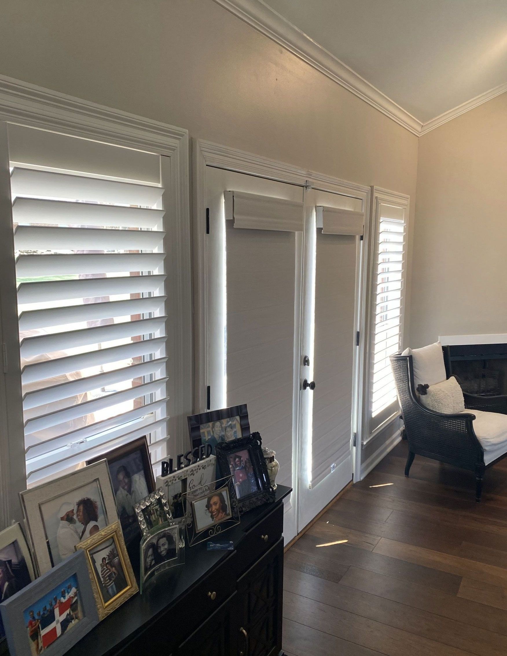 Room with white shutters, dark wood floors, and a black dresser displaying framed photos.