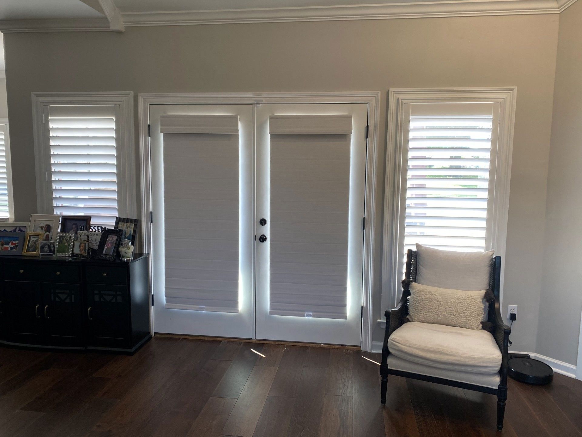 A room with dark wood floors, a set of double doors, shutters, and a black cabinet. A white armchair sits near the window.