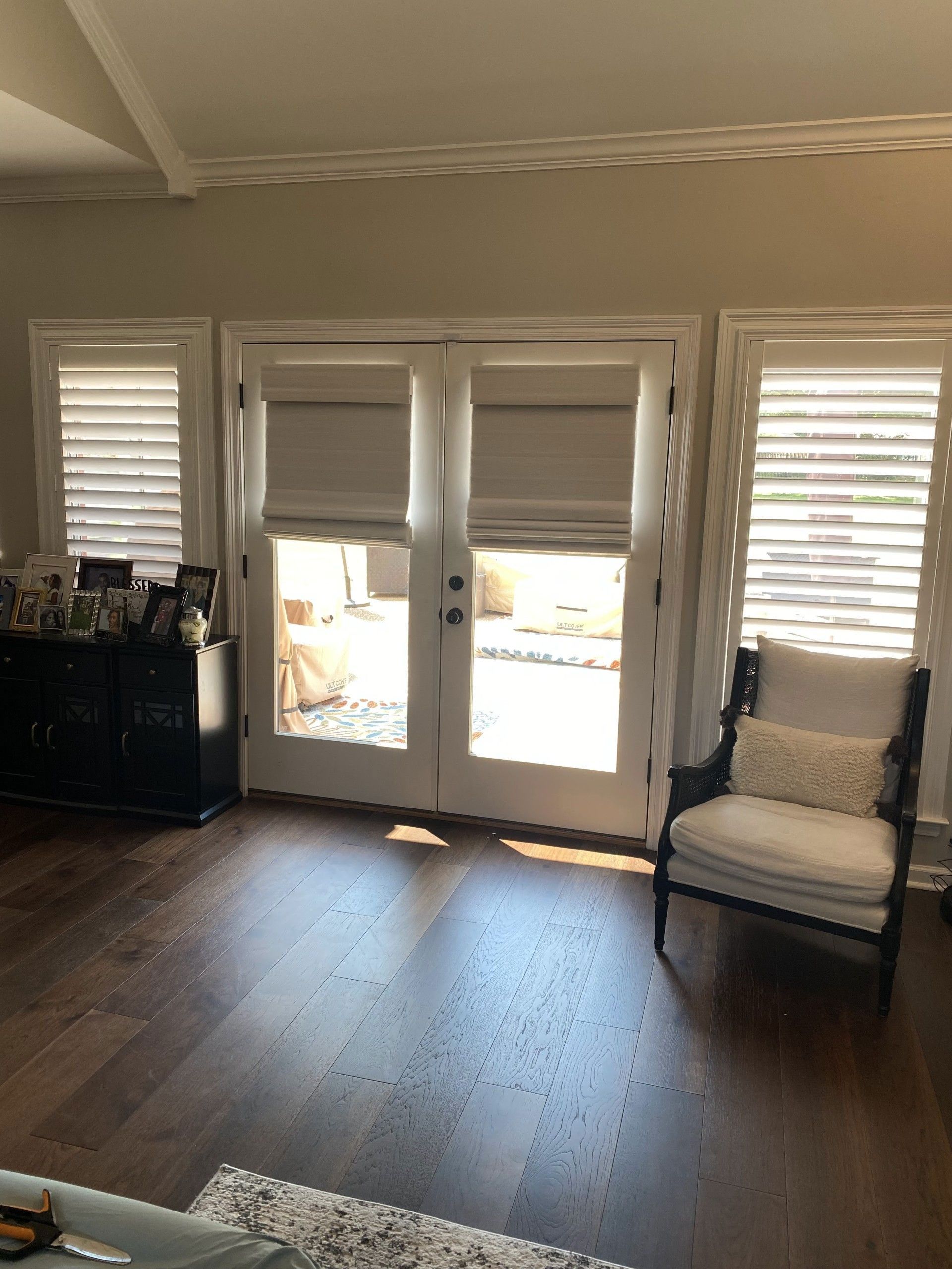 Living room with French doors flanked by shuttered windows. A dark cabinet and chair are on either side.