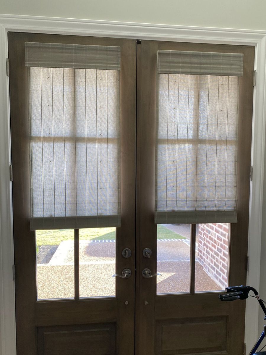 Two brown french doors with sheer, light-colored shades. Sunlight shines through the window panes.