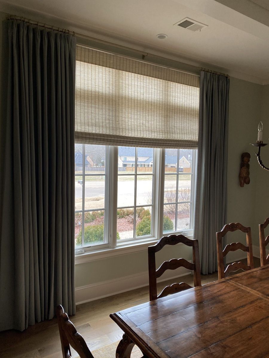 A window framed by gray curtains and a natural woven shade, with a dining table and chairs in front.