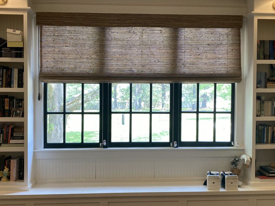A window with black frames and woven shade, flanked by bookshelves on a white built-in bench.