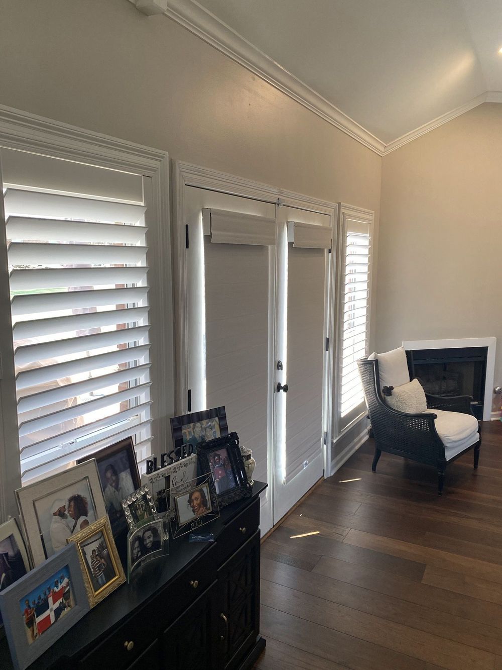A room with white shutters and doors, a fireplace, and a chair. A dark wooden dresser displays framed photos.