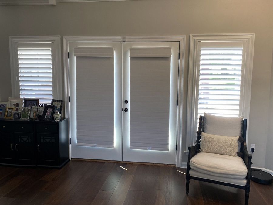 Room with closed white blinds and shutters. Two doors are flanked by windows, with furniture and a dark floor.