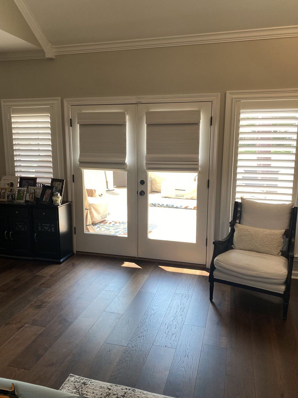 Interior view of a living room with French doors and two windows, all with window treatments. A black cabinet and chair sit on opposite sides of the doors.