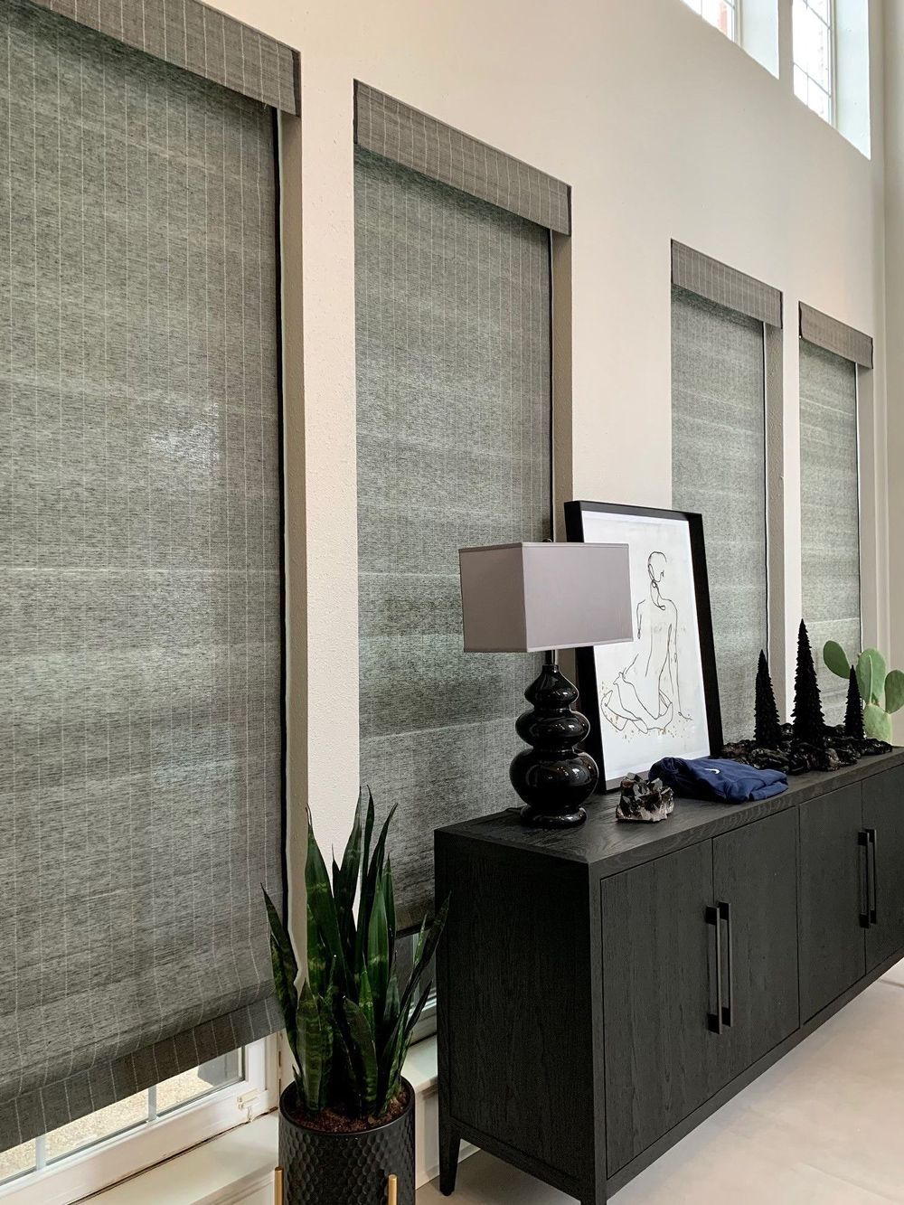 Interior with dark gray woven shades on tall windows. A black credenza with décor and a potted snake plant are in front.