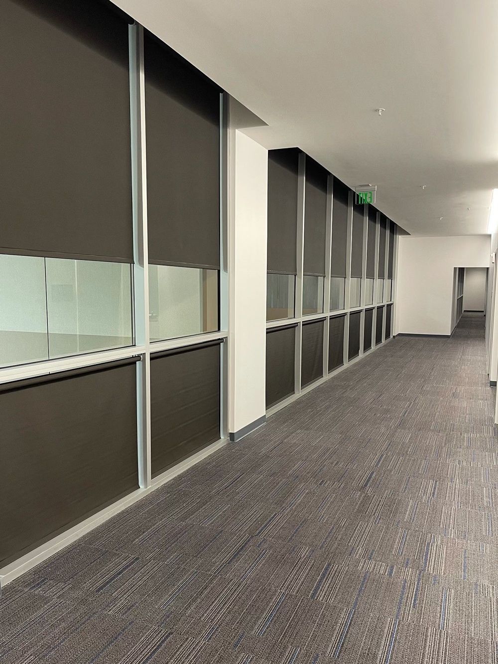 Dark hallway with large windows and dark shades. The floor is patterned carpet. There is an exit sign in the distance.