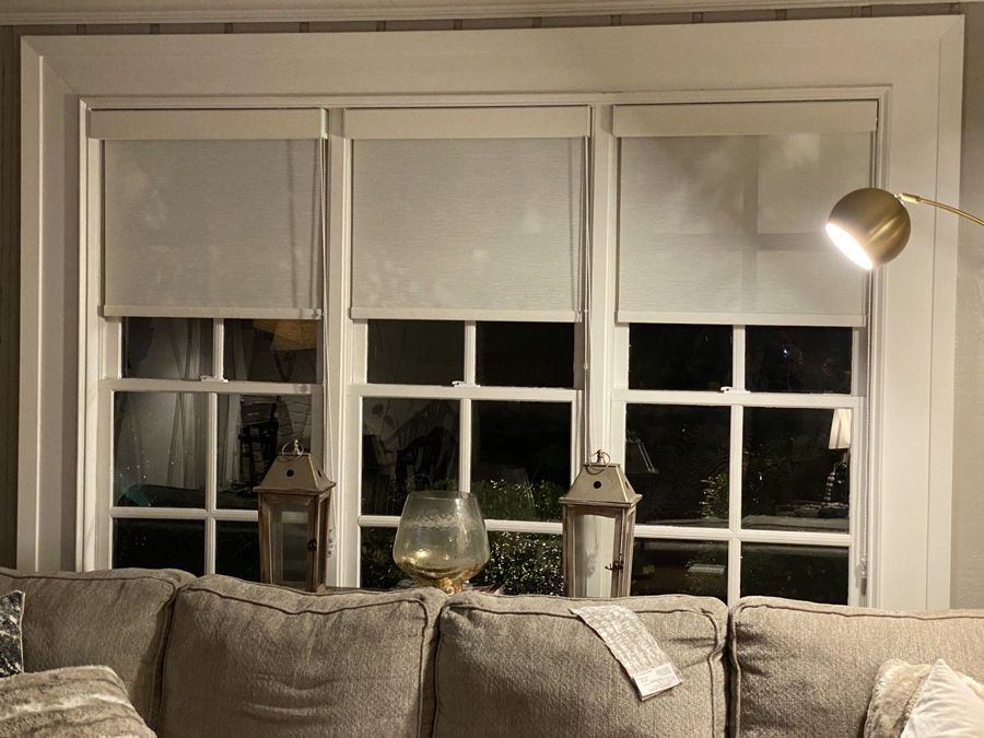 A lit window with three white blinds and a sectional sofa beneath. Decorative lanterns and a lamp are visible.