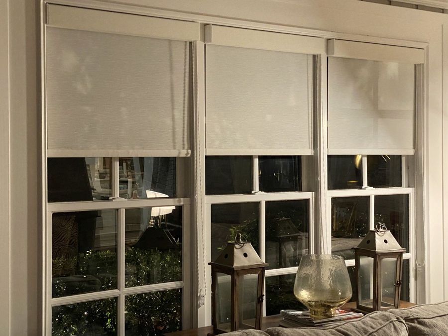 Three white-framed windows with light-colored roller shades partially covering the glass. Lanterns sit on a surface below.