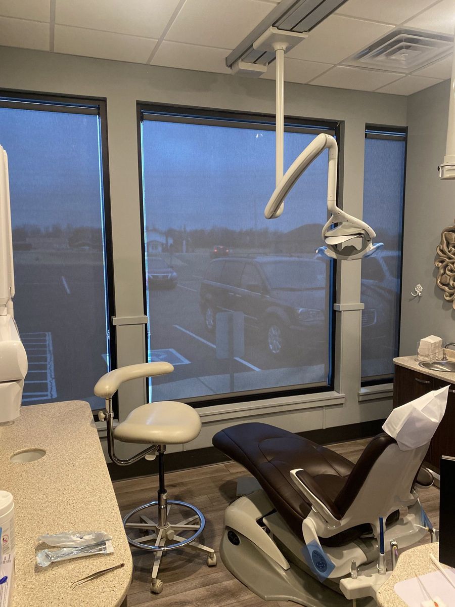 Dental office room with a patient chair, overhead lamp, and window with a roller blind.
